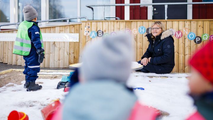 Alt-text: Barn leker utomhus i en snötäckt förskolegård medan en vuxen sitter och observerar.