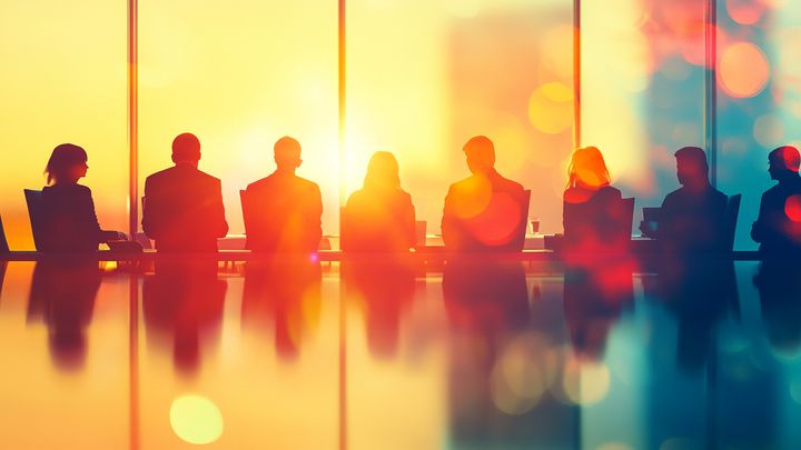 En grupp människor sitter vid ett konferensbord i ett modernt kontor, med ett stort fönster och soligt ljus i bakgrunden.