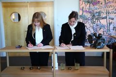 Två kvinnor skriver på dokument vid ett bord, omgiven av konstverk och dekorationer.