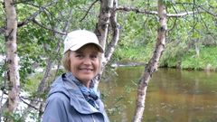 Kvinna i grå jacka och vit keps står framför en skogsbäck.