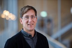 En person med glasögon och mörk tröja ler mot kameran. Bakgrunden är suddig med mjuka ljuskällor.