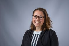 En leende person med glasögon och lockigt hår, klädd i en randig tröja och mörk kavaj, mot en grå bakgrund.