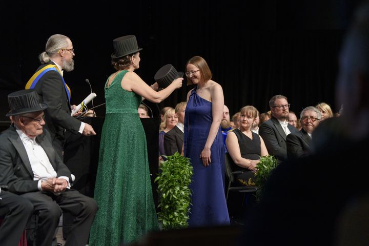 En person får en doktorshatt på en ceremoni, omgiven av andra i formella kläder.