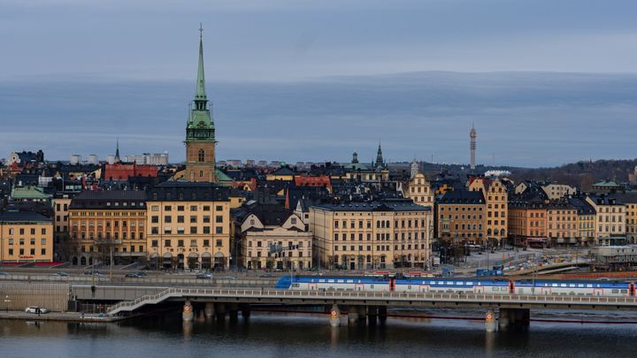 Panoramavy över en stad med historiska byggnader och en kyrka med ett högt torn. En bro sträcker sig över vattnet med ett tåg som passerar.