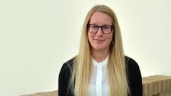En person med långt blont hår och glasögon som sitter vid en tegelvägg.