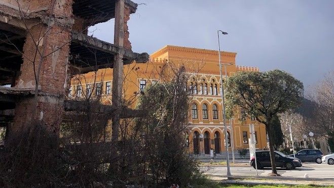 En ruinerad byggnad i Mostar från Bosnienkriget står i förgrunden, medan en ny, välbevarad byggnad syns i bakgrunden.