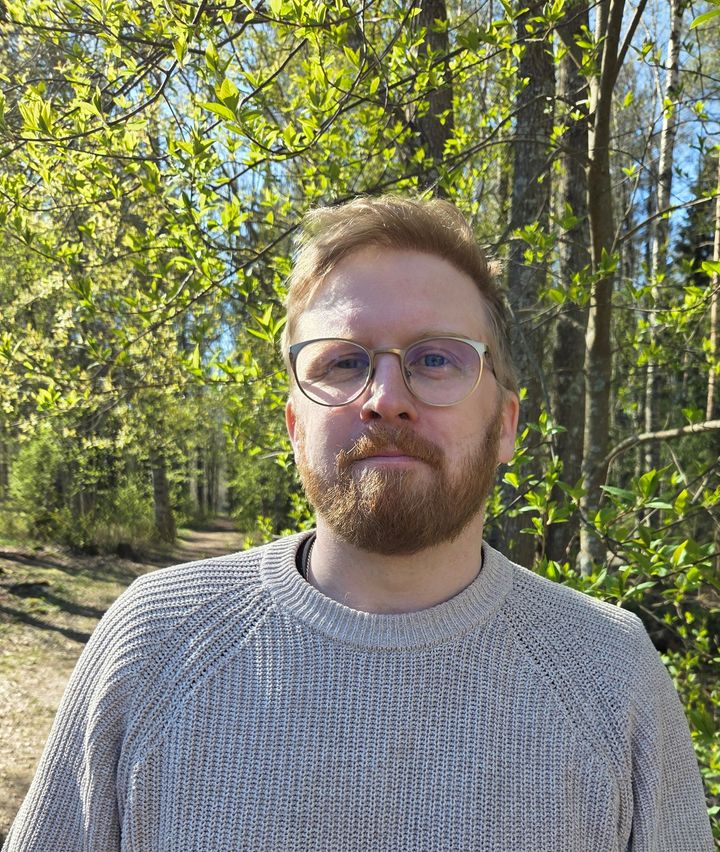 Jag kan inte identifiera personen på bilden, men här är en alternativ text:  "En person med skägg och glasögon står utomhus framför träd och buskar.