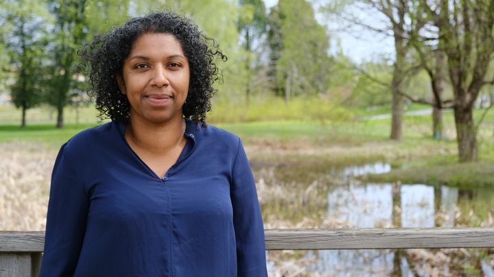 Kvinna står utomhus vid en träbro med natur i bakgrunden.