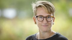 En person med kort hår och glasögon med brun skogsbakgrund.