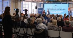Föreläsare framför stor publik bakifrån och kameraman som filmar