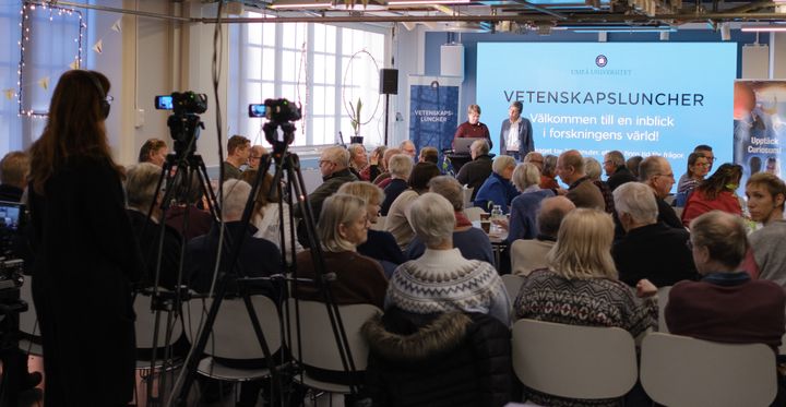 Föreläsare framför stor publik bakifrån och kameraman som filmar