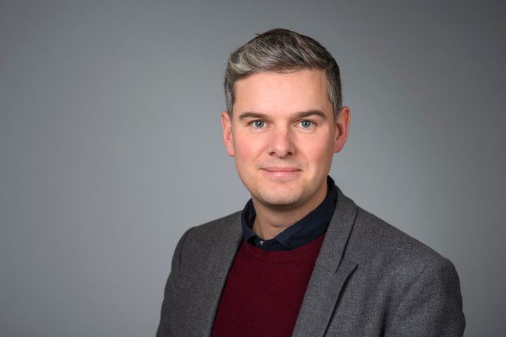 Man i grå kavaj och vinröd tröja står framför en grå bakgrund.
