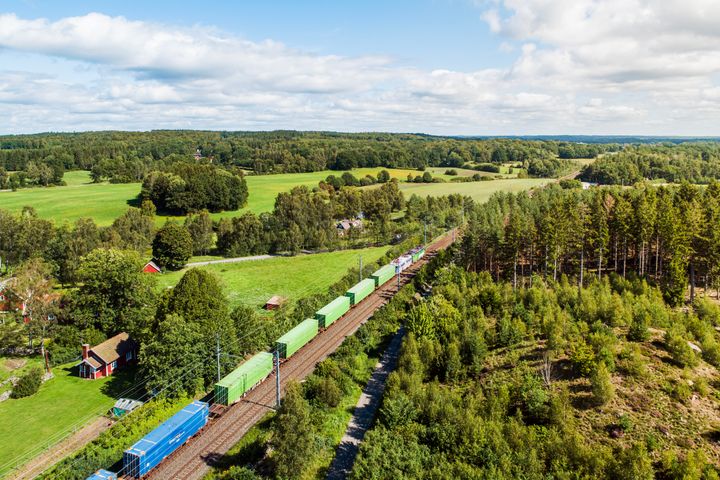Green Cargos godståg rullar genom vacker natur med åkrar och skog på sommaren.