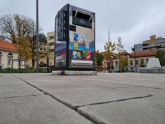 En Big belly-soptunna på ett torg i Borås, omgiven av byggnader och träd.