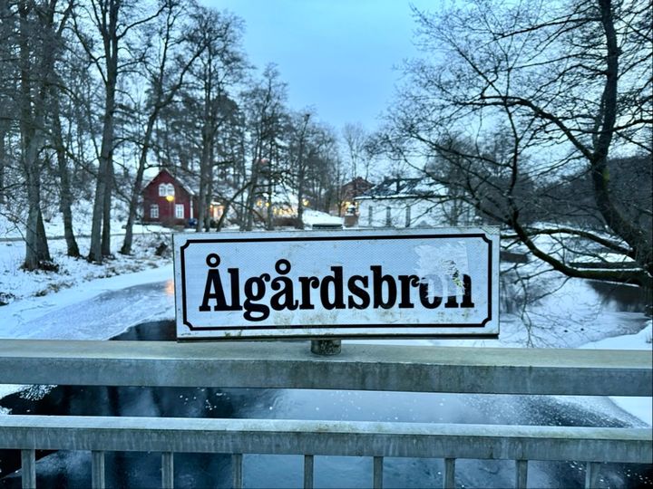 Skylt med texten "Ålgårdsbron" på en bro, omgiven av vinterlandskap med snö och träd.