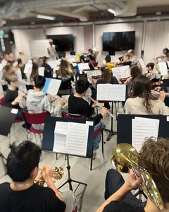 En grupp orkestermusiker som spelar tillsammans i ett repetitionsrum. Olika instrument som brass och träblås ses i förgrunden.