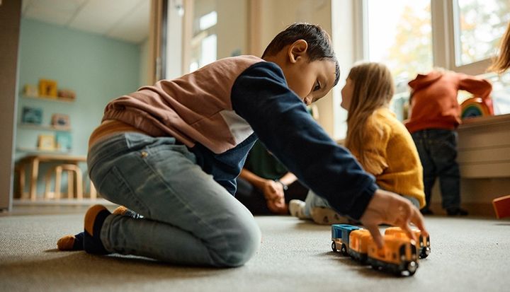Ett barn leker med ett leksakståg på golvet i ett klassrum, medan andra barn sitter i bakgrunden.