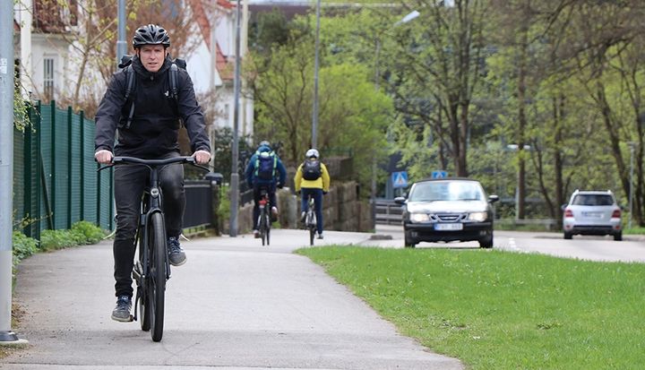 Person cyklar på en gång- och cykelväg med fler människor och bilar i bakgrunden.