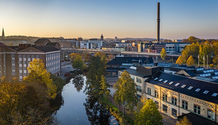 Vy över en stad med en flod, byggnader och en hög skorsten. Trädens lövverk skiftar i höstfärger.
