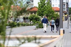 Tre personer promenerar på en trädbevuxen gågata i en stad, med byggnader och grönska i bakgrunden.