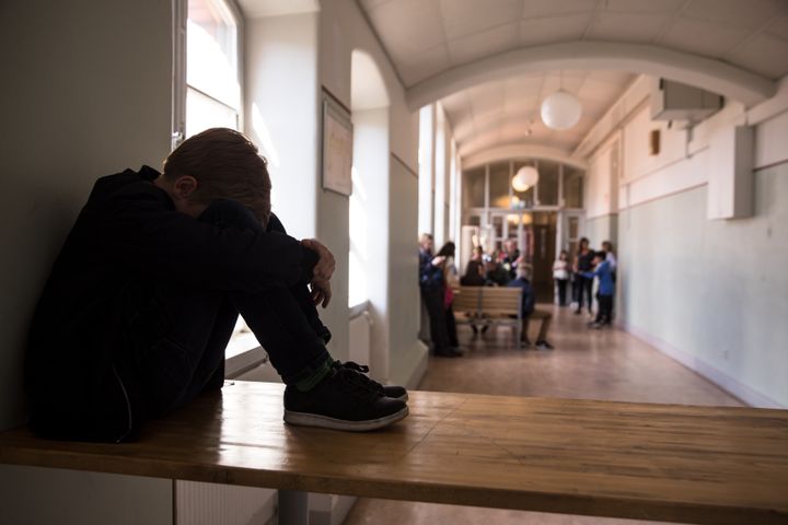 En person sitter ihopkrupen på en bänk i en skolkorridor, medan en grupp människor står samlade längre bort.