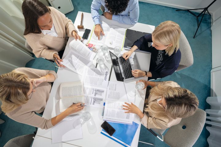 Fem personer diskuterar runt ett mötesbord med papper och laptop.