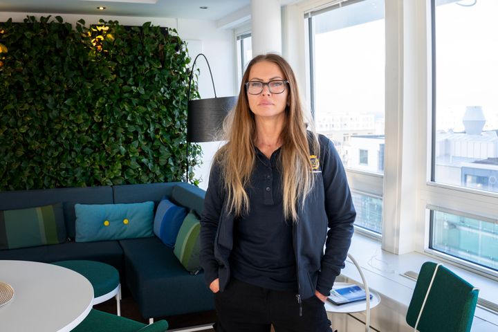 Kvinna med långt hår och glasögon står i ett modernt kontor med grön vägg i bakgrunden.