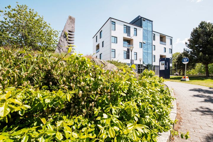 Ljust flerfamiljshus med grönska i förgrunden under klar himmel.