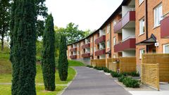 Hyresfastighet med röd tegelfasad, något svängd huskropp. Gångväg framför fastigheten och till vänster är gräsmatta samt enar.