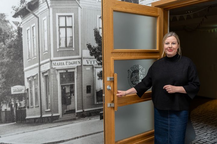 Kvinna står vid en öppen dörr, leende. Bakgrund med gammaldags fasad.