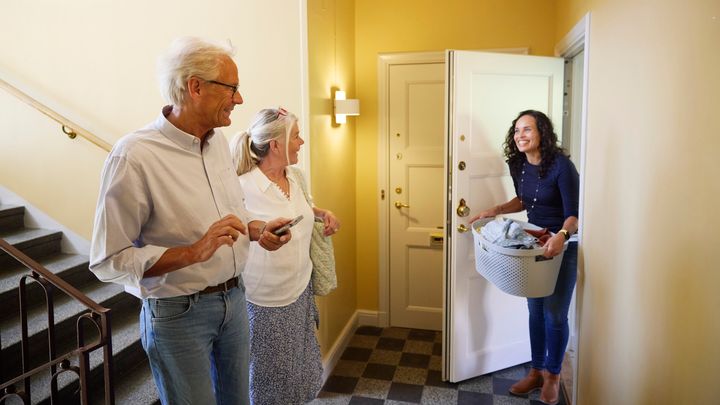 Kvinna kömmer ut från öppen dörr, ler mot äldre par som går i trappan i trapphuset.