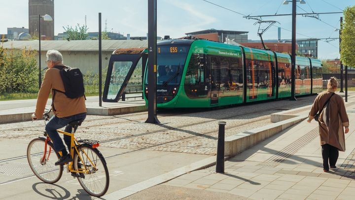 Man cyklar över spåren till spårvagnen. En spårvagn står på stationen och en kvinna promenerar längs spåren.