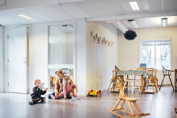 Aikuinen%20ja%20kaksi%20lasta%20leikkim%E4ss%E4%20lattialla%20valoisassa%20p%E4iv%E4kotitilassa%2C%20taustalla%20p%F6yti%E4%20ja%20tuoleja.