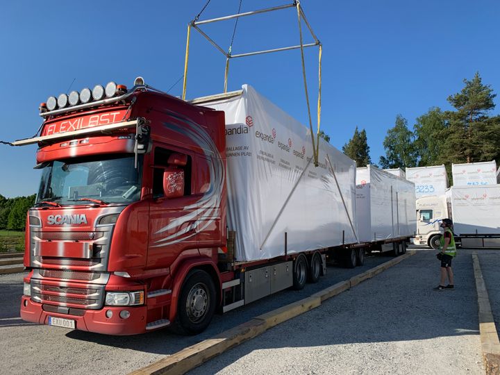Punainen%20rekka%20kuljettaa%20muovisuojattuja%20Expandian%20moduuleita%20ty%F6maalle%20samalla%2C%20kun%20nosturi%20valmistelee%20moduulin%20nostoa%20paikoilleen.%20Ty%F6ntekij%E4%20seuraa%20asennusta%20aurinkoisella%20ty%F6maa-alueella.