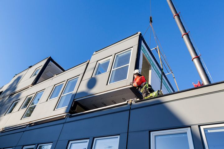 Ty%F6ntekij%E4%20ohjaa%20nosturin%20avulla%20moduulin%20asennusta%20kaksikerroksisen%20rakennuksen%20katolla%20kirkkaan%20sinisen%20taivaan%20alla.%20Moduulirakennuksen%20suuret%20ikkunat%20ja%20moderni%20julkisivu%20n%E4kyv%E4t%20kuvassa%20selke%E4sti.