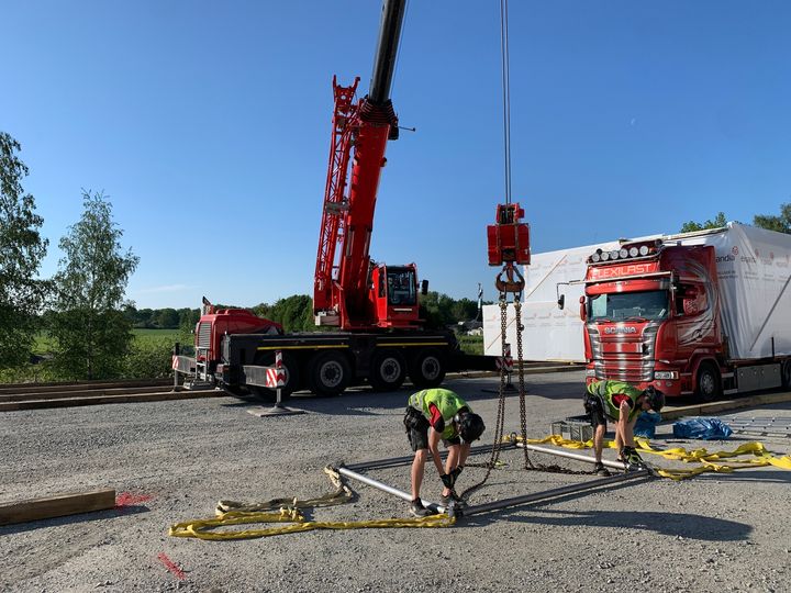 Ty%F6ntekij%F6it%E4%20valmistelemassa%20nostokalustoa%20moduulien%20siirtoa%20varten%20ty%F6maalla.%20Kuvassa%20n%E4kyy%20punainen%20nosturi%2C%20moduuleja%20kuljettava%20rekka%20sek%E4%20ty%F6ntekij%F6it%E4%20kiinnitt%E4m%E4ss%E4%20nostokehikkoa%20sorapintaisella%20alueella%20aurinkoisessa%20s%E4%E4ss%E4.