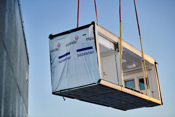 Ilmassa%20nostettava%20moduulielementti%20ty%F6maalla%20kirkasta%20taivasta%20vasten.%20Kuvassa%20n%E4kyy%20keskener%E4inen%20moduuli%2C%20jonka%20sis%E4tilat%2C%20ikkunat%20ja%20kattorakenteet%20ovat%20jo%20valmiina%20ennen%20paikalleen%20asennusta.%20Punaiset%20nostoliinat%20pit%E4v%E4t%20moduulia%20ilmassa%20asennuksen%20aikana.
