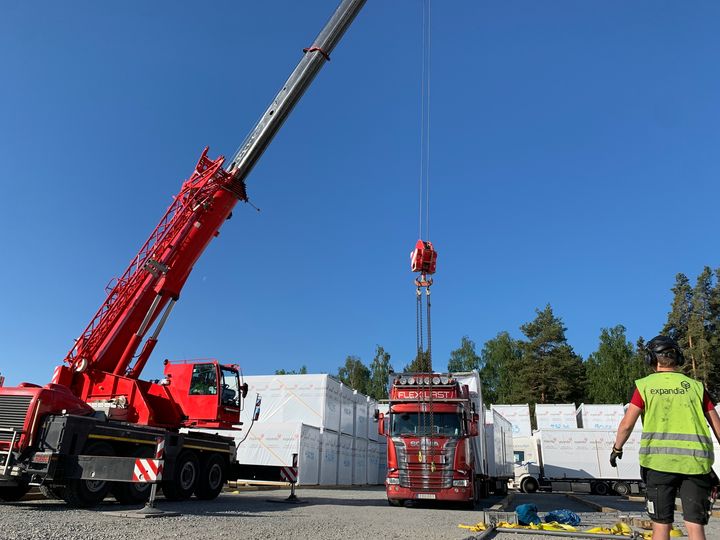 Punainen%20mobiilinosturi%20ty%F6skentelee%20moduulivarastointialueella%20moduulielementtien%20siirron%20aikana.%20Kuvassa%20n%E4kyy%20nostokalustoa%2C%20moduulielementtej%E4%20kuljettava%20rekka%20sek%E4%20ty%F6ntekij%E4%20valvomassa%20asennusty%F6t%E4%20kirkkaassa%20kes%E4s%E4%E4ss%E4.