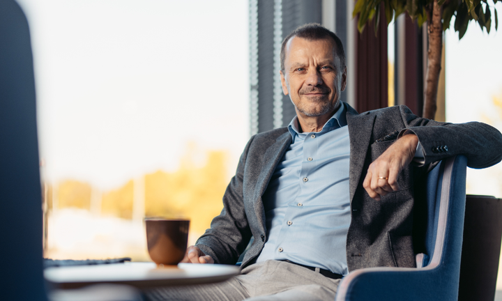 En man sitter i en stol bredvid ett fönster med en kaffekopp på bordet intill.