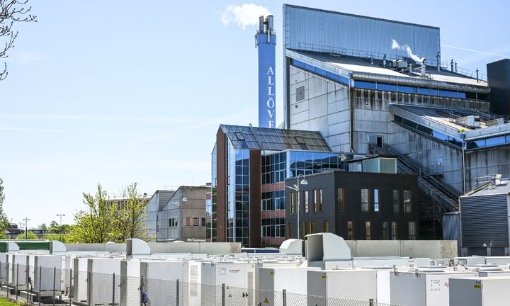 Bild av en batteripark med flera vita batteriskåp. Fjärrvärmeverket, Allöverket, i bakgrunden.