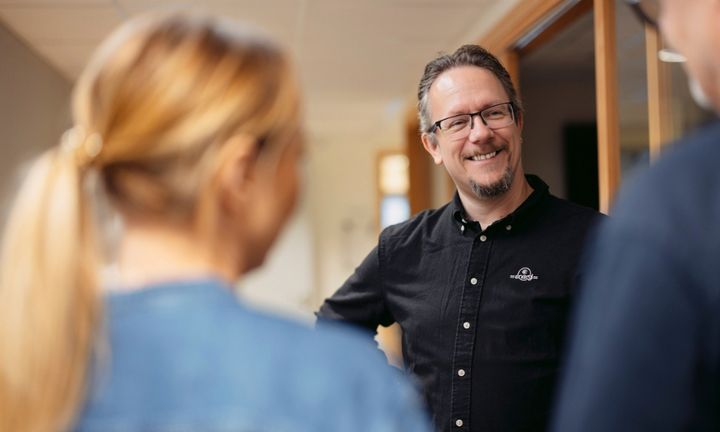 En man i glasögon och svart skjorta står och pratar med två personer, som är delvis i bild.