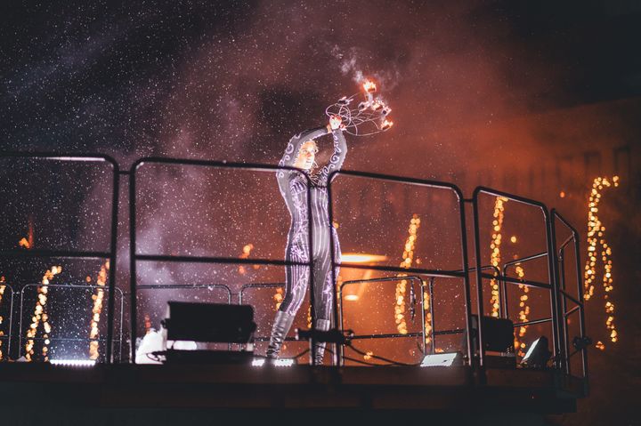 En person klädd i glittrande dräkt jonglerar med eldslungor på en upphöjd plattform, med gnistor som sprider sig i luften.