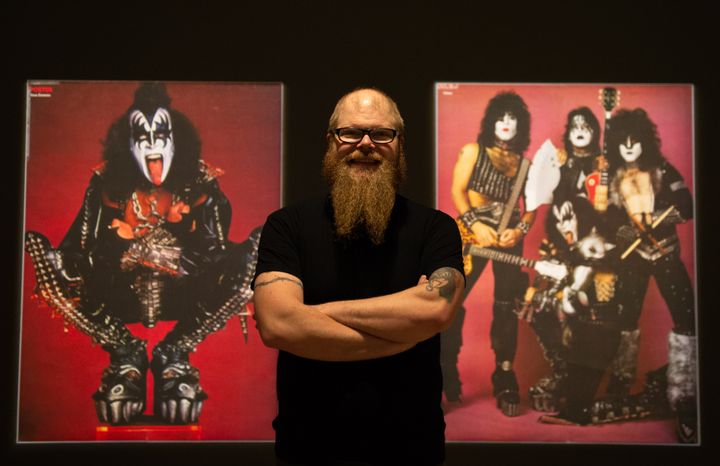 En man med skägg står framför två stora affischer av rockbandet KISS i kostym och smink.