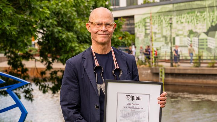 En person står utomhus vid en å och håller ett diplom.