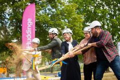 Fem personer med spadar som kastar sand, i bakgrunden syns en rosa flagga med texten Huddinge.