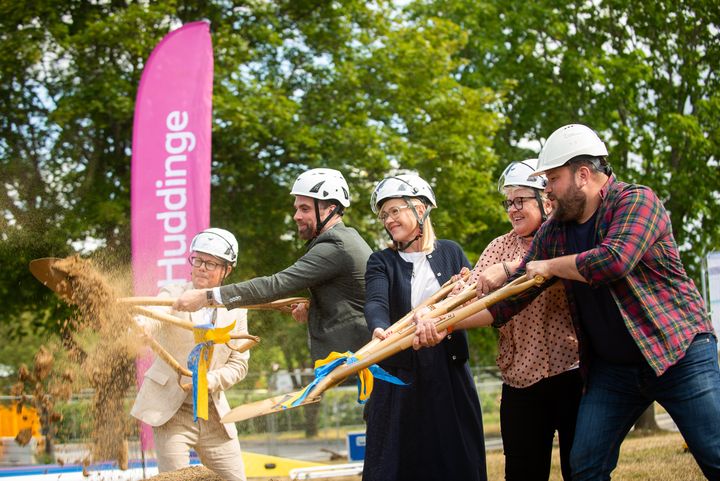 Fem personer med spadar som kastar sand, i bakgrunden syns en rosa flagga med texten Huddinge.