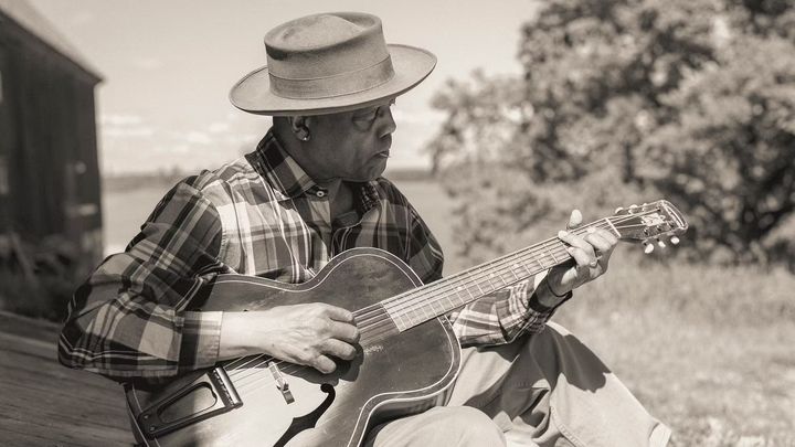 En person sitter utomhus och spelar akustisk gitarr.