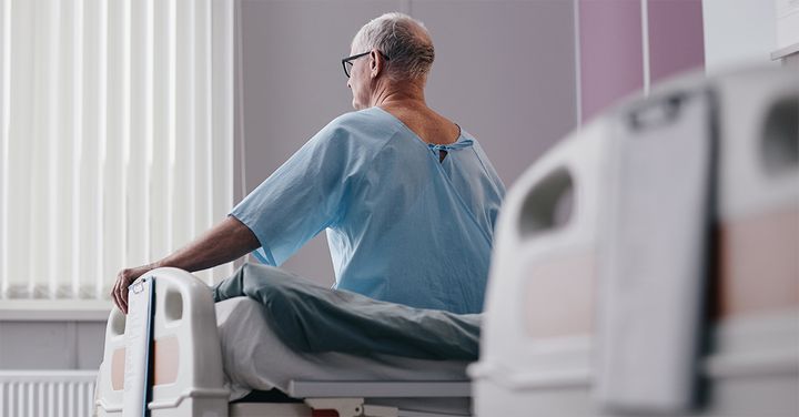 En äldre man i sjukhuskläder sitter på en sjukhussäng och tittar bort.