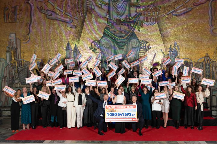 En stor grupp finklädda, glada människor står på en scen med stora checkar i händerna.