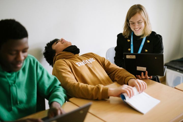En glad lärare och två elever som arbetar vid ett bord med datorer.